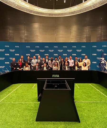 A group of people gathered around a table, engaged in discussion in front of a large presentation screen.