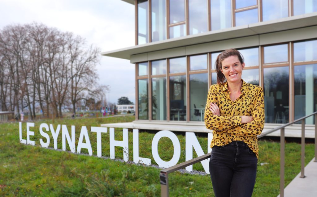 Lea Sprunger, Swiss Track and Field athlete standing in front of the Le Synahtlon, AISTS building. Wearing yellow dotted shirt and black jeans.