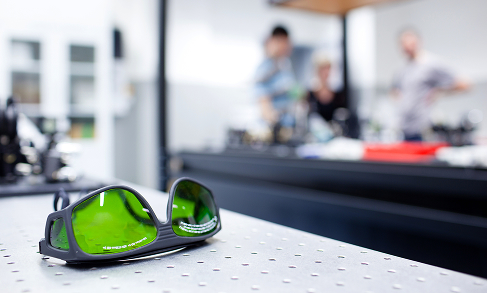 Green safety glasses on a lab table with researchers blurred in the background representing the equipment testing services of AISTS