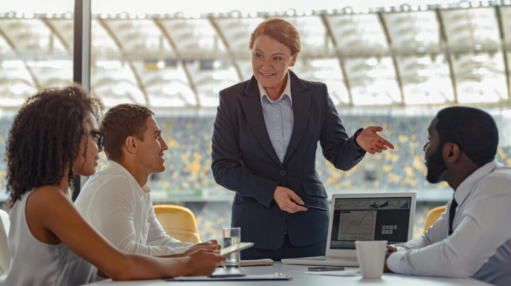 A sports manager leading a meeting with a diverse team of 4 sports managers in a stadium conference room, discussing performance charts on a laptop. 
