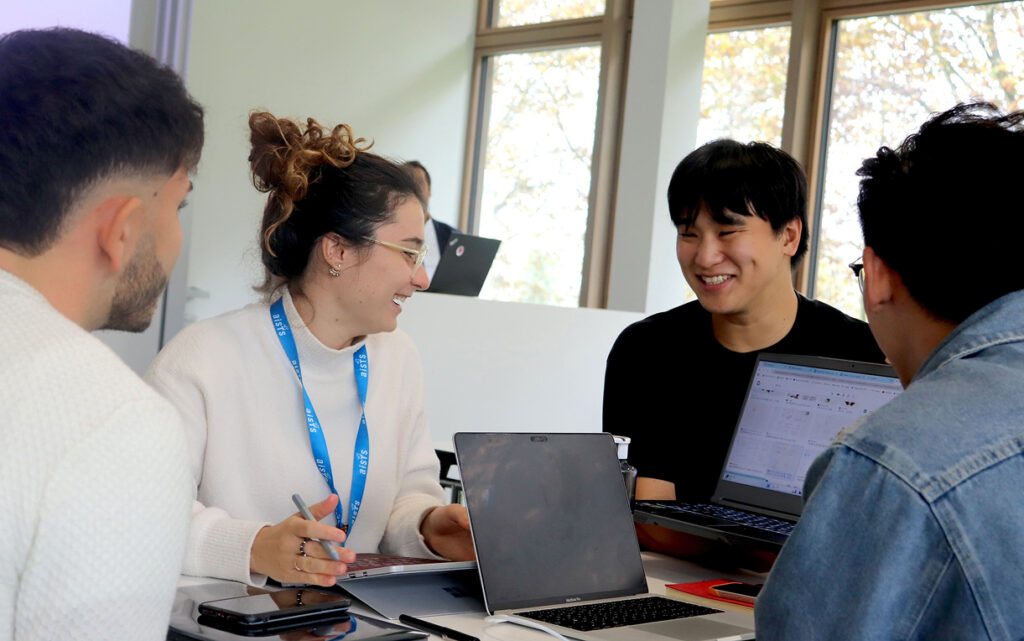 Four AISTS students pursuing a sports management degree collaborating around a table in a bright classroom, using laptops and notebooks during a group project, smiling and discussing ideas.