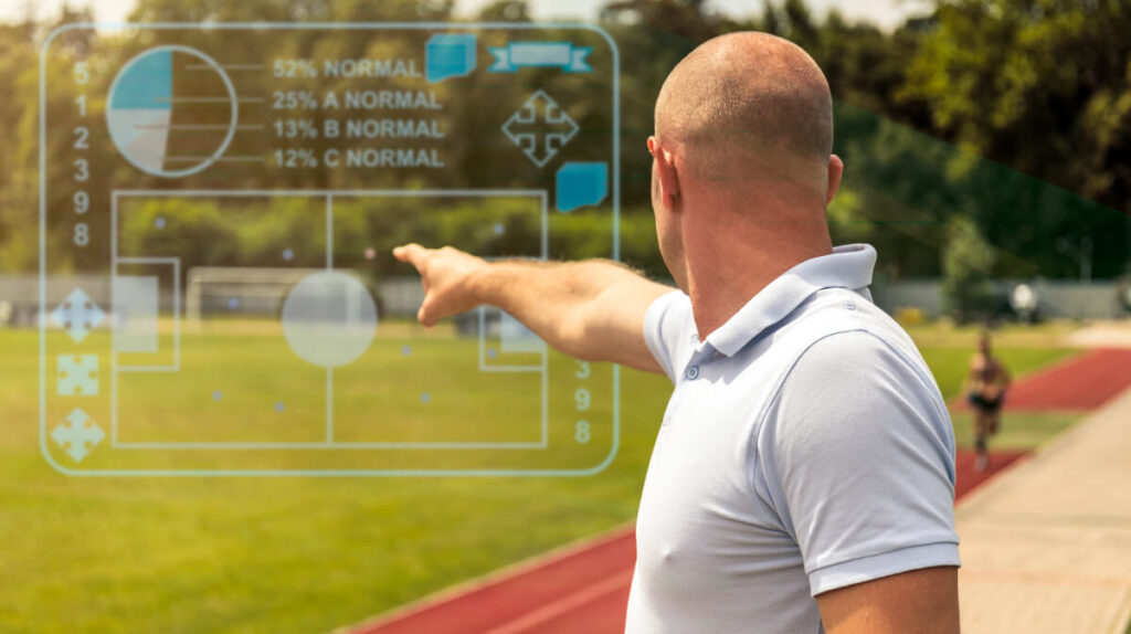 Coach analyzing player statistics with augmented reality sports technology on a football field, with an athlete running in the background.